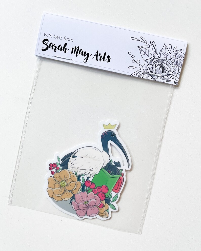 May include: A sticker featuring a white ibis wearing a gold crown, holding a green bin with black bags inside. The ibis is surrounded by pink, red, and yellow flowers.
