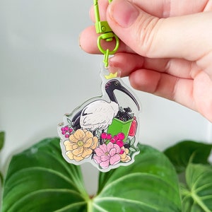 以下が含まれることがあります： 白い頭と頭に冠をかぶった白いトキが、緑と赤の花柄の上に立っています。トキは赤い取っ手のついた緑色の袋を持っています。デザインは、緑色の金属製の留め具が付いた透明なアクリルキーホルダーに施されています。