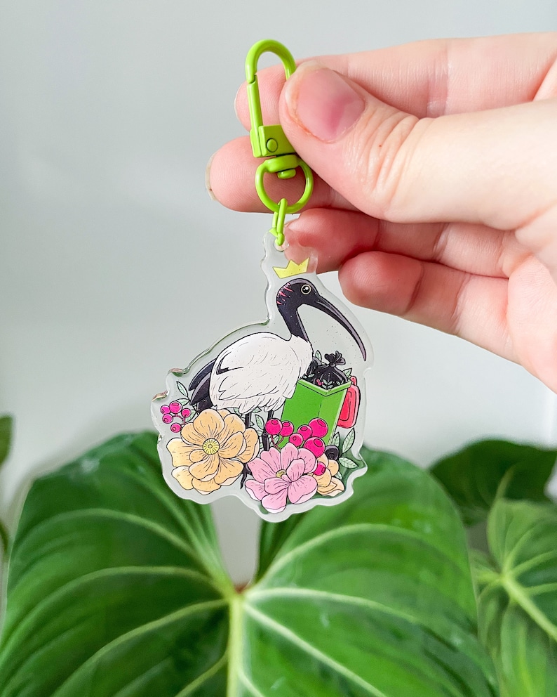 以下が含まれることがあります： 白い頭と頭に冠をかぶった白いトキが、緑と赤の花柄の上に立っています。トキは赤い取っ手のついた緑色の袋を持っています。デザインは、緑色の金属製の留め具が付いた透明なアクリルキーホルダーに施されています。