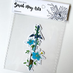 May include: A sticker featuring a blue and green sword with white flowers wrapped around it. The sticker is on a white background with a small heart and the text "with love, from Sarah May Arts".