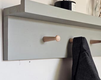 May include: A grey wooden wall shelf with six wooden pegs. A dark grey jacket hangs from one peg. The shelf is mounted on a white wall. Decorative items sit on the shelf above.
