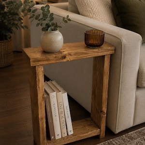 May include: A wooden side table with a natural finish. It has a lower shelf holding three books and a top surface with a small vase of greenery and a brown glass bowl. The table is next to a beige sofa.