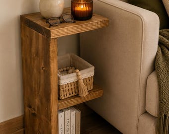 May include: A wooden side table with two shelves. The top shelf features a small vase with greenery, spectacles, and a lit candle in a glass holder. The lower shelves display a woven basket and books. The table is next to a beige sofa.