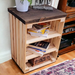 May include: A wooden shelf unit with four shelves, made from natural wood with a dark brown stained top. The shelf unit is filled with books and a decorative wooden box. A silver metal sculpture of a group of people sitting on a bench is on top of the shelf unit.