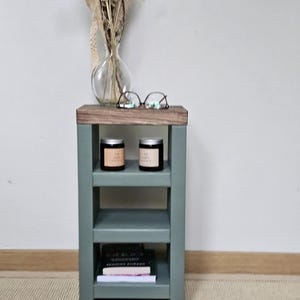 May include: A sage green wooden side table with three shelves and a dark wood top. Eyeglasses rest on the top. Two candles in black jars and books are on the lower shelves. A clear vase with dried flowers is behind the table.