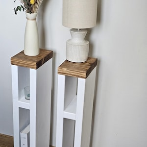 May include: Two white wooden side tables with a brown wood top. Each table has two shelves and a small opening at the bottom. The table on the left has a book titled "KINFOLK" on the shelf. The table on the right has a small vase with sticks in it on the shelf.