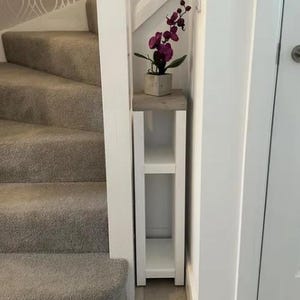 May include: A slim, white wooden shelf unit with two shelves and a grey top. A small potted plant with purple flowers sits on top of the shelf. The unit is placed in a hallway next to a staircase with grey carpet.