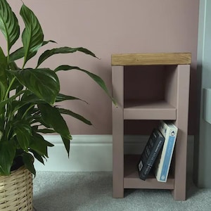 May include: A small, dusky pink wooden side table with two shelves and a light oak top. Two books are placed on the lower shelf. A large green plant in a woven basket is to the left of the table.