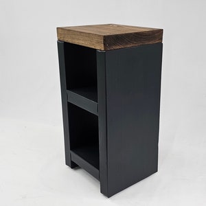 May include: A small, dark gray wooden side table with two shelves. The top is a contrasting brown wood. The table is rectangular and has a simple, modern design, suitable for a living room or bedroom.