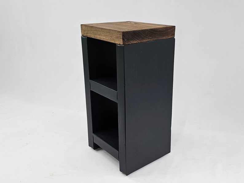 May include: A small, dark gray wooden side table with two shelves. The top is a contrasting brown wood. The table is rectangular and has a simple, modern design, suitable for a living room or bedroom.