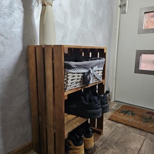 May include: A wooden crate shoe rack with three shelves. The top shelf has a wicker basket with a grey and white checked liner. The middle shelf has two pairs of black trainers. The bottom shelf has two pairs of brown hiking boots. The shoe rack is in a hallway with a door and a rug.