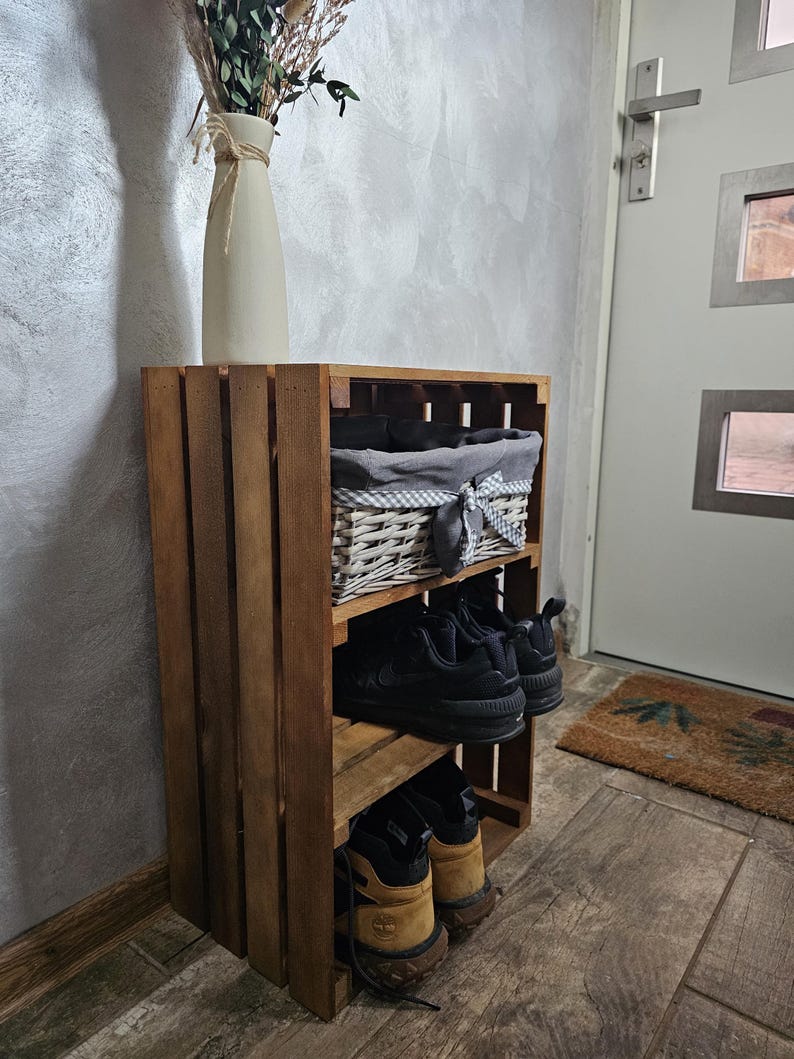 May include: A wooden crate shoe rack with three shelves. The top shelf has a wicker basket with a grey and white checked liner. The middle shelf has two pairs of black trainers. The bottom shelf has two pairs of brown hiking boots. The shoe rack is in a hallway with a door and a rug.