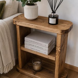 May include: A rustic, wooden side table with two shelves. The top shelf holds a white potted plant and a black diffuser. The middle shelf displays three books: "THE KINFOLK", "LIVE BEAUTIFULLY", and "THIS IS HOME". A candle sits on the bottom shelf.