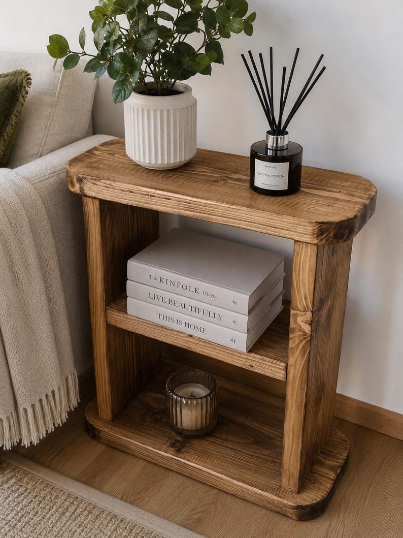 May include: A rustic, wooden side table with two shelves. The top shelf holds a white potted plant and a black diffuser. The middle shelf displays three books: "THE KINFOLK", "LIVE BEAUTIFULLY", and "THIS IS HOME". A candle sits on the bottom shelf.
