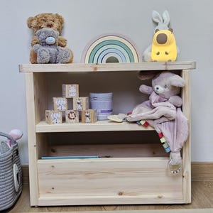 May include: A light-coloured wooden shelf unit with two shelves and a drawer. The top shelf holds a teddy bear, a rainbow-shaped wooden toy, and a yellow night light. The middle shelf has wooden blocks and a plush toy. A grey basket is on the left.