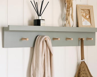 May include: A grey wall-mounted shelf unit with five wooden pegs. A beige scarf and a brown net bag hang from the pegs. The shelf holds a black diffuser, a vase with dried plants, and a framed picture. The wall is white with vertical panelling.
