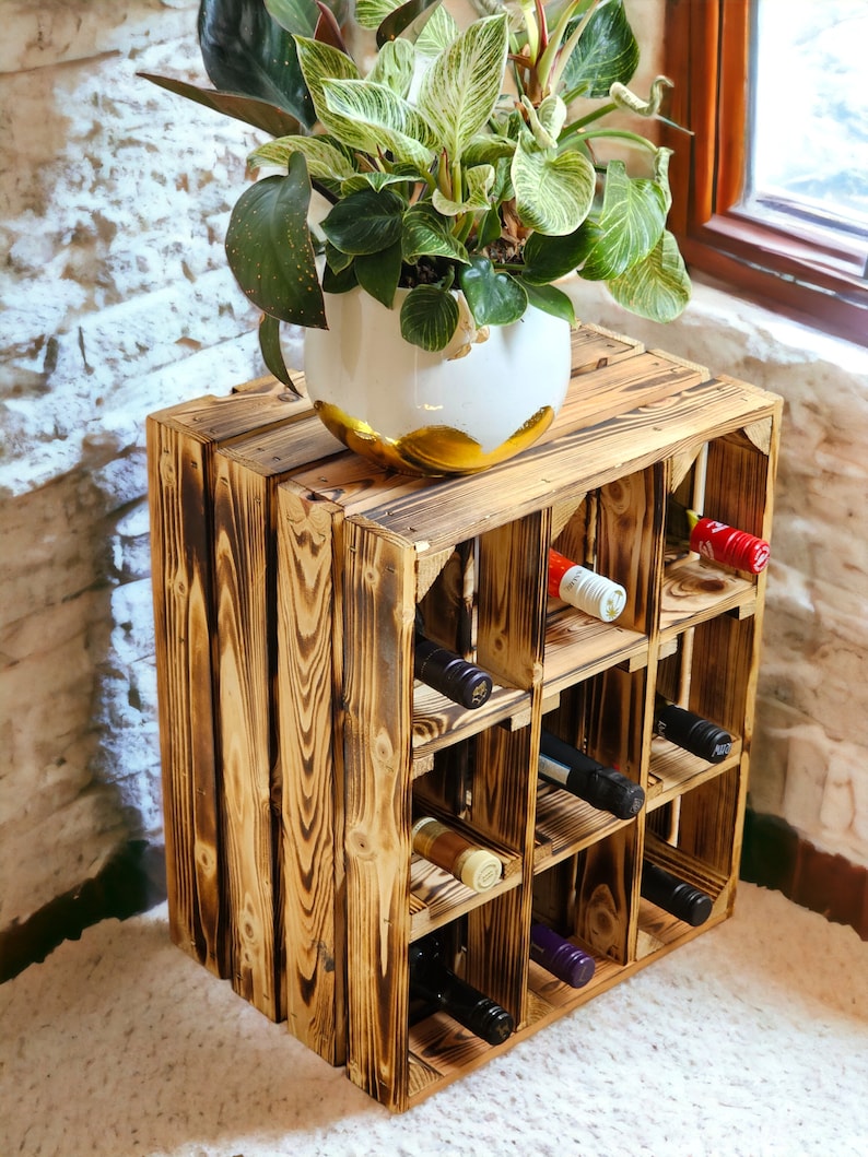 May include: A rustic wooden wine rack with a dark, charred finish, holding several wine bottles. The rack is topped with a white and gold ceramic planter with a green and white leafy plant. The rack is made of wooden crates.