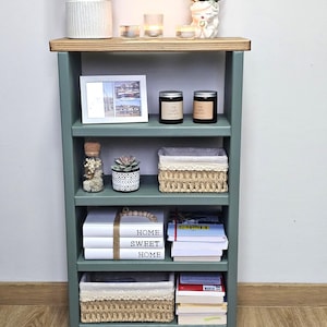 May include: A green wooden bookshelf with four shelves. The top shelf has a white lamp, two candles, and a white ceramic planter with dried flowers. The second shelf has a framed photo, two candles, and a small glass jar with dried flowers. The third shelf has a small succulent planter, a woven basket, and a glass jar with dried flowers. The fourth shelf has a stack of books with the words "Home Sweet Home" on the top book, a woven basket, and a stack of books. The bookshelf is standing on a wooden floor.
