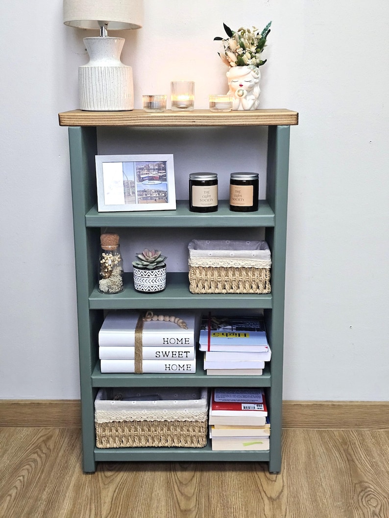 May include: A green wooden bookshelf with four shelves. The top shelf has a white lamp, two candles, and a white ceramic planter with dried flowers. The second shelf has a framed photo, two candles, and a small glass jar with dried flowers. The third shelf has a small succulent planter, a woven basket, and a glass jar with dried flowers. The fourth shelf has a stack of books with the words "Home Sweet Home" on the top book, a woven basket, and a stack of books. The bookshelf is standing on a wooden floor.