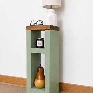 May include: A sage green side table with a dark wood top. A white lamp with a beige lampshade sits on top. A pair of black glasses, a candle, and a wooden pear are displayed on the table. The table is 30 inches tall.