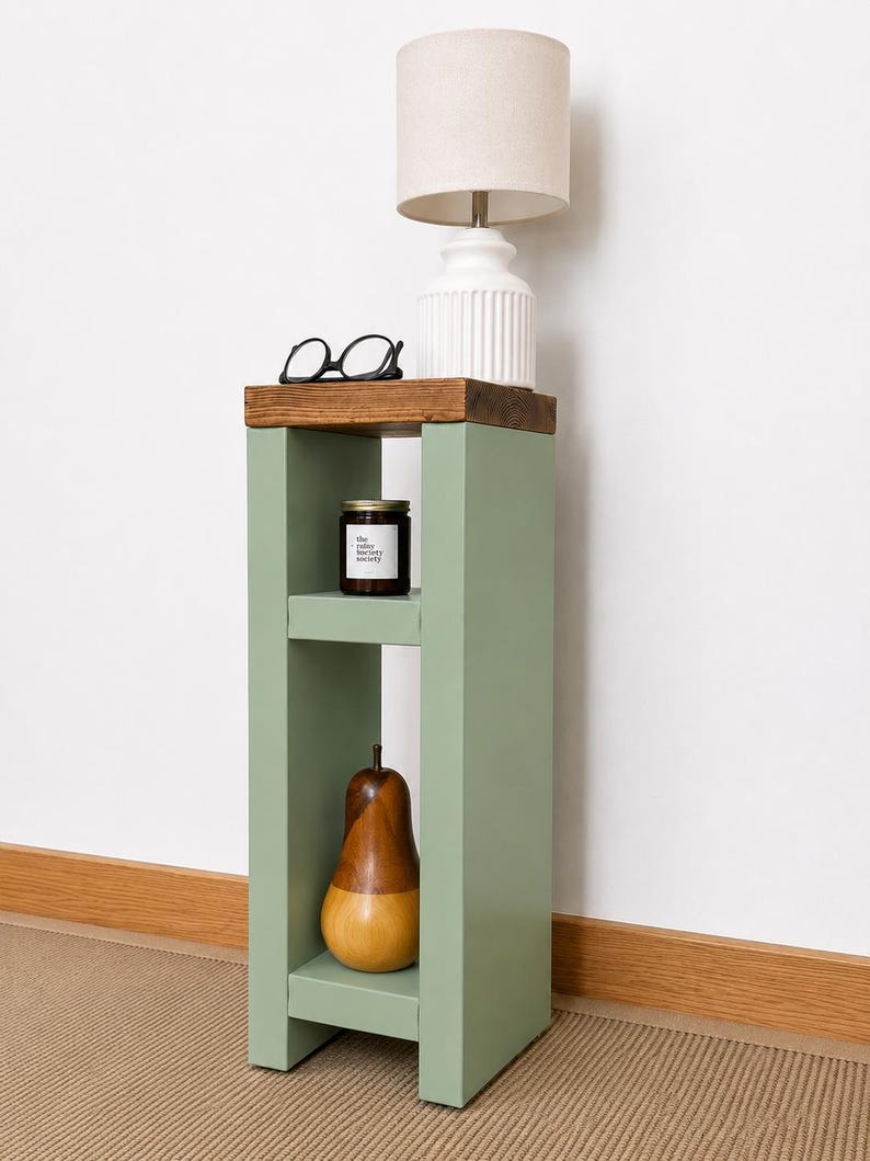 May include: A sage green side table with a dark wood top. A white lamp with a beige lampshade sits on top. A pair of black glasses, a candle, and a wooden pear are displayed on the table. The table is 30 inches tall.