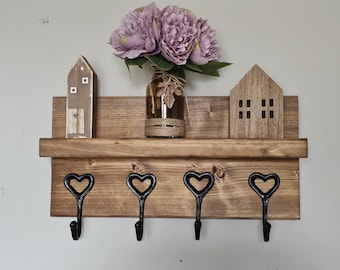 May include: Wooden wall mounted shelf with three black heart-shaped hooks. The shelf has two small wooden houses on either side and a vase with purple flowers in the center.