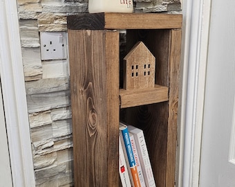 May include: A wooden shelf unit with two shelves, a small wooden house figurine, and several books. The shelf is stained a dark brown color and has a rustic look. The shelf is sitting on a wooden floor.