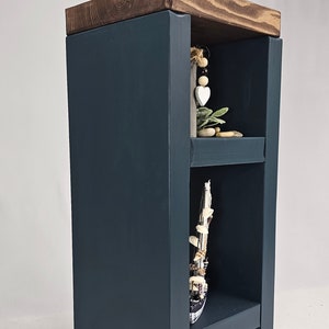 May include: A dark blue wooden side table with two shelves and a dark wood top. The top shelf holds a cream-colored vase with light blue and green flowers. Decorative items are displayed on the shelves.