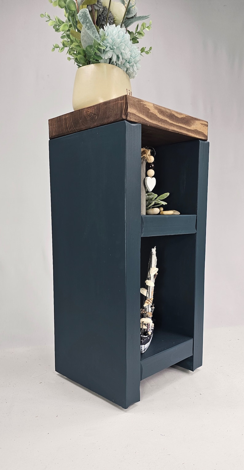 May include: A dark blue wooden side table with two shelves and a dark wood top. The top shelf holds a cream-colored vase with light blue and green flowers. Decorative items are displayed on the shelves.
