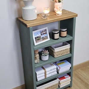 A wooden painted bookshelf with candles, framed photos, two baskets, decorative jars, and stacked books  placed on a wooden floor against a white wall.