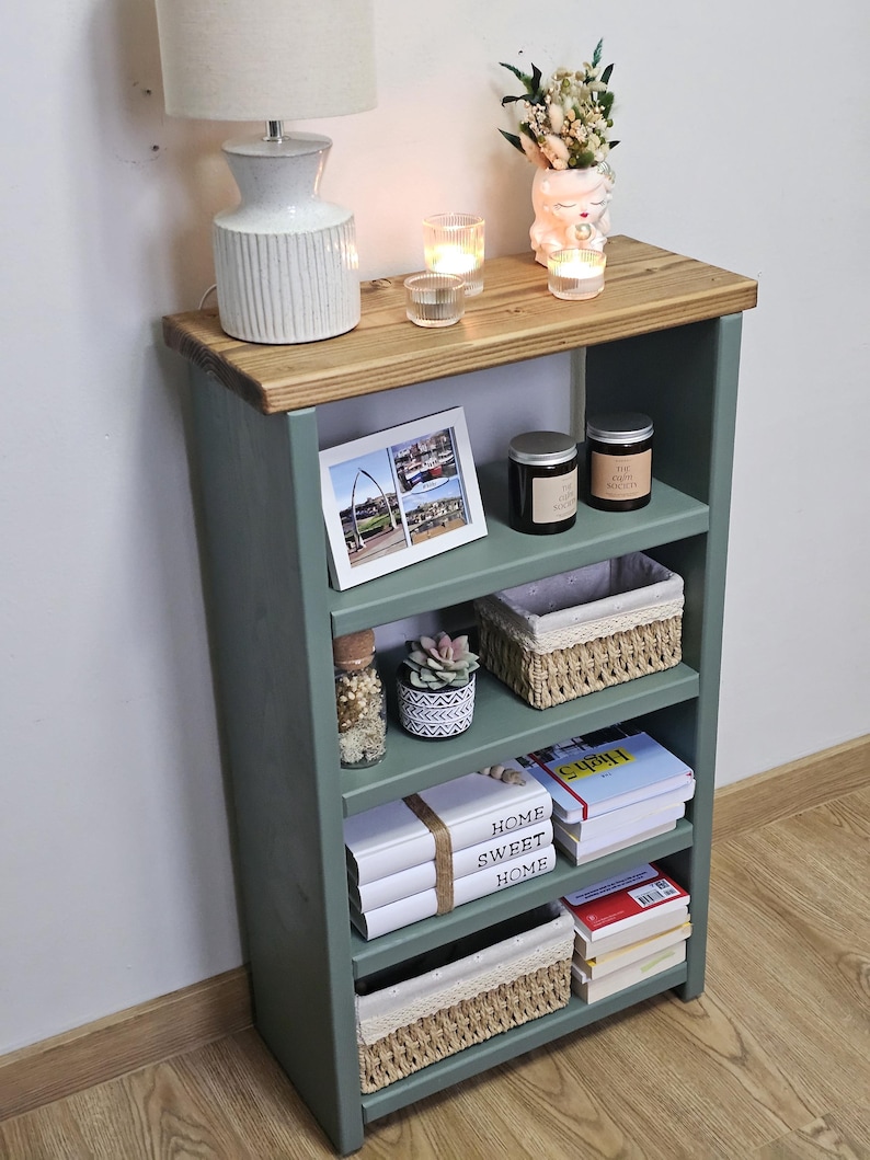 A wooden painted bookshelf with candles, framed photos, two baskets, decorative jars, and stacked books  placed on a wooden floor against a white wall.