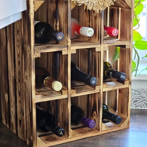 May include: A rustic, wooden wine rack with nine compartments, holding various wine bottles. The rack has a charred wood finish, giving it a vintage look. A woven mat sits atop the rack, and a plant is visible in the background.