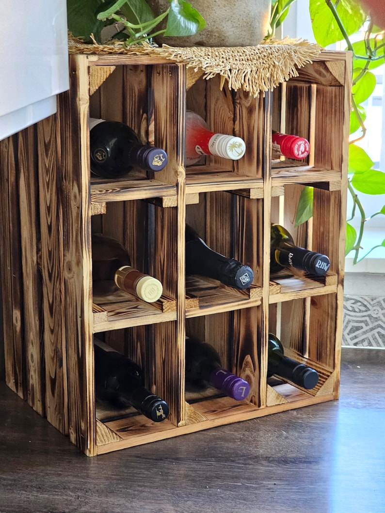 May include: A rustic, wooden wine rack with nine compartments, holding various wine bottles. The rack has a charred wood finish, giving it a vintage look. A woven mat sits atop the rack, and a plant is visible in the background.