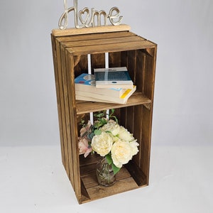 May include: A wooden crate shelf with two shelves, a metal "Home" sign on top, and a bouquet of white and pink flowers in a glass vase on the bottom shelf. The shelf is filled with books.