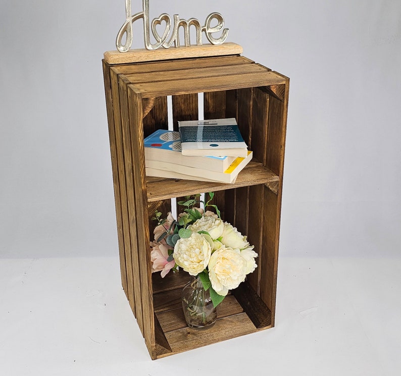 May include: A wooden crate shelf with two shelves, a metal "Home" sign on top, and a bouquet of white and pink flowers in a glass vase on the bottom shelf. The shelf is filled with books.