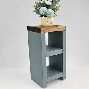 May include: A small, wooden side table painted in a muted blue-gray. The top is a contrasting dark brown, supporting a small vase with light blue and white flowers. The table has two shelves and a simple, modern design.
