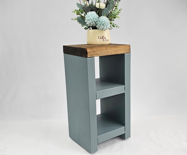 May include: A small, wooden side table painted in a muted blue-gray. The top is a contrasting dark brown, supporting a small vase with light blue and white flowers. The table has two shelves and a simple, modern design.
