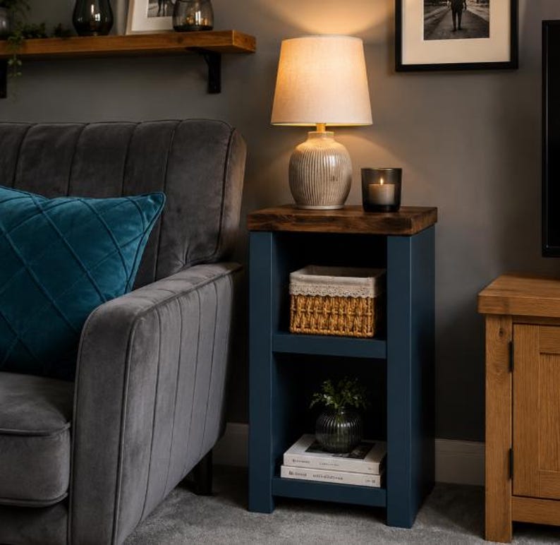 May include: A navy blue wooden side table with a dark brown top. The table has two shelves, one with a woven basket and the other with books and a small plant. A lamp with a beige shade sits on top.