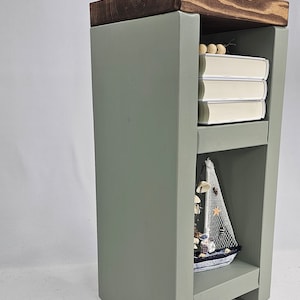 May include: A sage green wooden side table with a dark stained top. The table has two shelves, one with books and the other with a decorative sailboat. A vase with flowers sits on top. The table is approximately 24 inches tall.