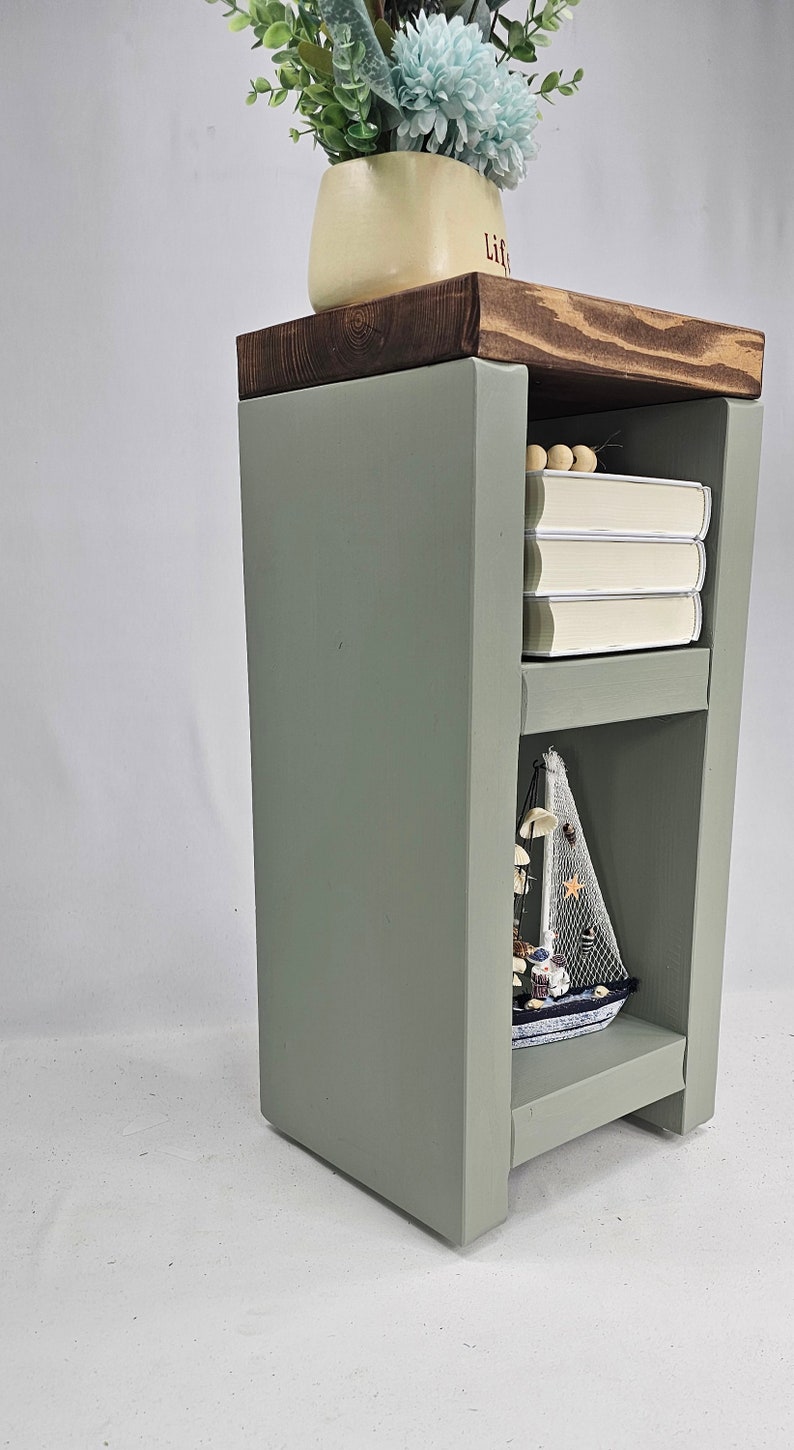 May include: A sage green wooden side table with a dark stained top. The table has two shelves, one with books and the other with a decorative sailboat. A vase with flowers sits on top. The table is approximately 24 inches tall.