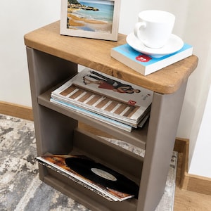 May include: A small side table in brown and grey with a wooden top. A framed photo, a white teacup and saucer, and a book are on top. Magazines and vinyl records are stored on the shelves below. The photo frame has the word "Scarborough" on it.