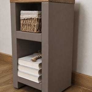 May include: A gray nightstand with a natural wood top. The nightstand has two shelves, one with a woven basket and the other with three books. A decorative item sits on top. The nightstand is on a wooden floor.