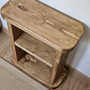 May include: A small, wooden, two-tiered shelf with a natural finish. The shelf has rounded corners and a lower shelf for storage. The wood grain is visible, and the piece is sturdy and simple in design. It is suitable for a variety of uses.