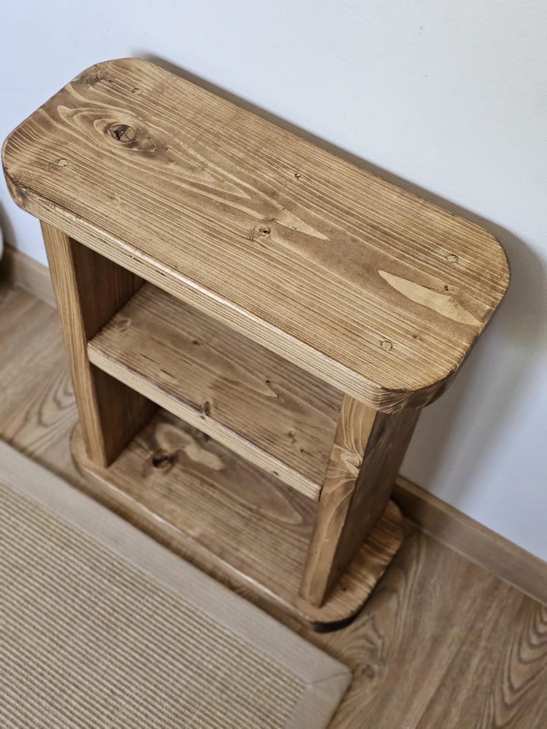 May include: A small, wooden, two-tiered shelf with a natural finish. The shelf has rounded corners and a lower shelf for storage. The wood grain is visible, and the piece is sturdy and simple in design. It is suitable for a variety of uses.