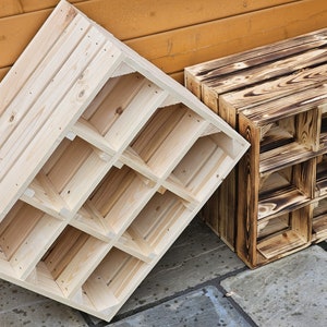 May include: Two wooden wine racks. One is unfinished, light-colored wood, and the other is a darker, burnt-wood finish. Each rack has multiple square compartments for storing bottles. The racks are angled against a wooden wall and a stone surface.