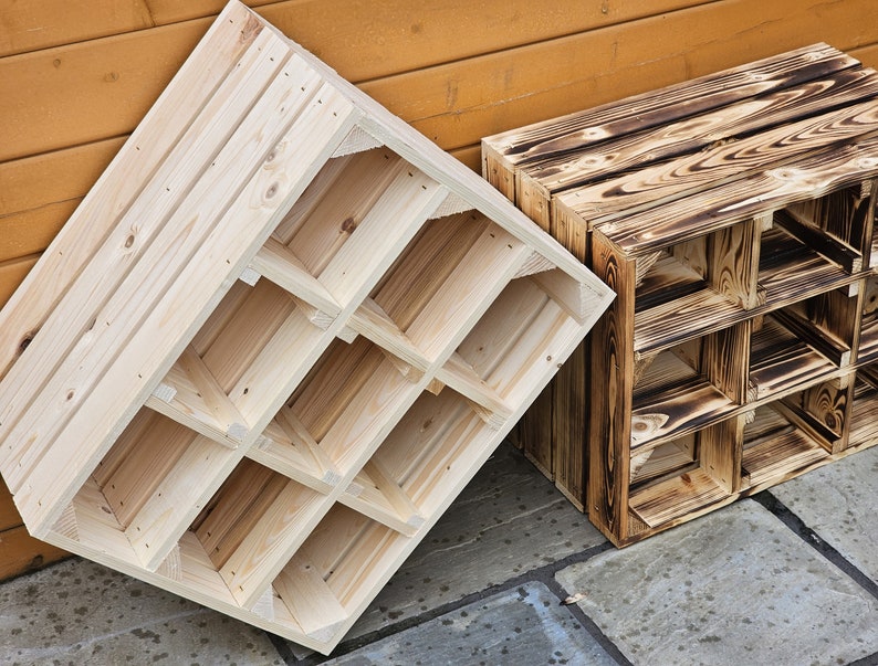 May include: Two wooden wine racks. One is unfinished, light-colored wood, and the other is a darker, burnt-wood finish. Each rack has multiple square compartments for storing bottles. The racks are angled against a wooden wall and a stone surface.