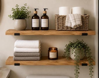 May include: Two natural wood floating shelves with black metal brackets. The top shelf holds a potted green plant, two amber glass pump bottles labeled Hand Wash and Lotion, and a woven basket with two toilet paper rolls and a plaid towel.