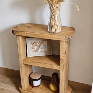May include: A rustic, wooden side table with two shelves. A clear glass vase filled with dried wheat stalks sits on top. A framed picture, a candle, and a wooden pear-shaped object are on the shelves. The table is made of light brown wood.