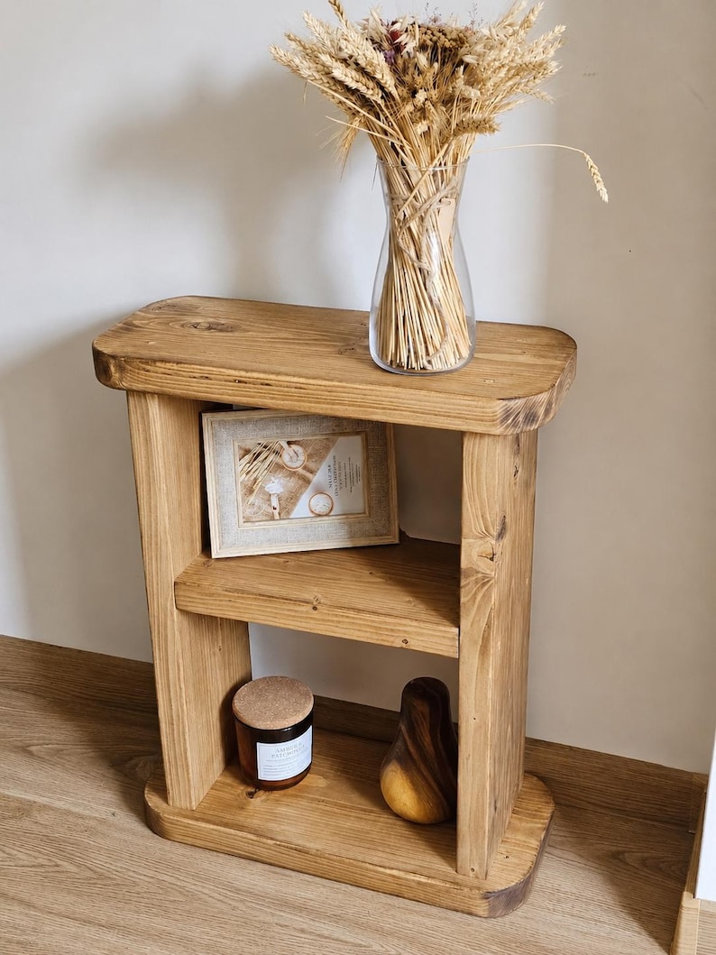 May include: A rustic, wooden side table with two shelves. A clear glass vase filled with dried wheat stalks sits on top. A framed picture, a candle, and a wooden pear-shaped object are on the shelves. The table is made of light brown wood.