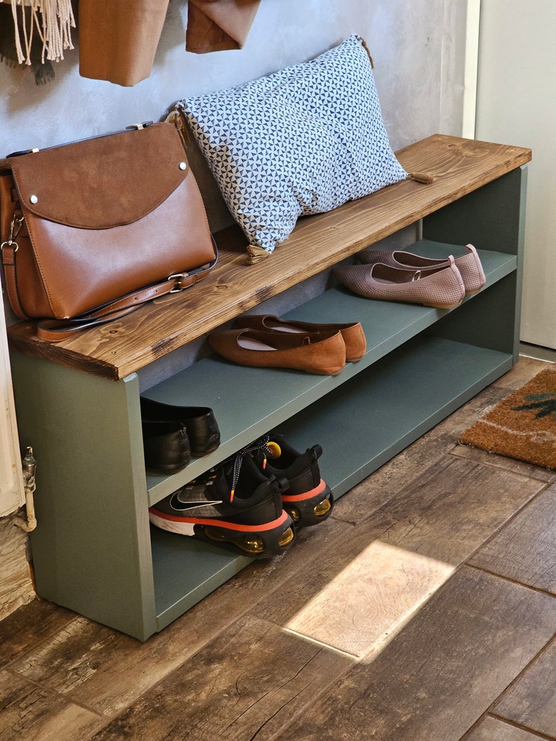 May include: A green wooden shoe rack with two shelves. The top shelf has a brown wooden top and a brown leather purse. A blue and white patterned pillow is on the top shelf. The bottom shelf has a pair of black and orange sneakers and a pair of brown flats.